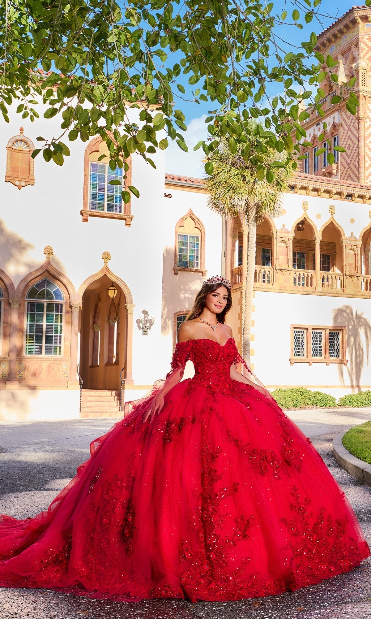 A young woman wearing the Quinceanera Dress PR30116 By Princesa poses outside a grand building with arched windows and a balcony on a sunny day.